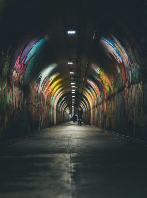 Zwei Menschen gehen durch einen leicht beleuchteten mit Graffiti besprühten Tunnel. Es soll widerspiegeln, dass sich Menschen mit Morbus Crohn oft sozial isolieren.
