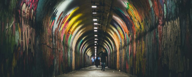 Zwei Menschen gehen durch einen leicht beleuchteten mit Graffiti besprühten Tunnel. Es soll widerspiegeln, dass sich Menschen mit Morbus Crohn oft sozial isolieren.
